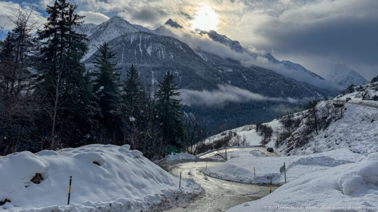 Narrow icy road between deep snowbanks descending toward forested mountains under a glowing cloudy sky.