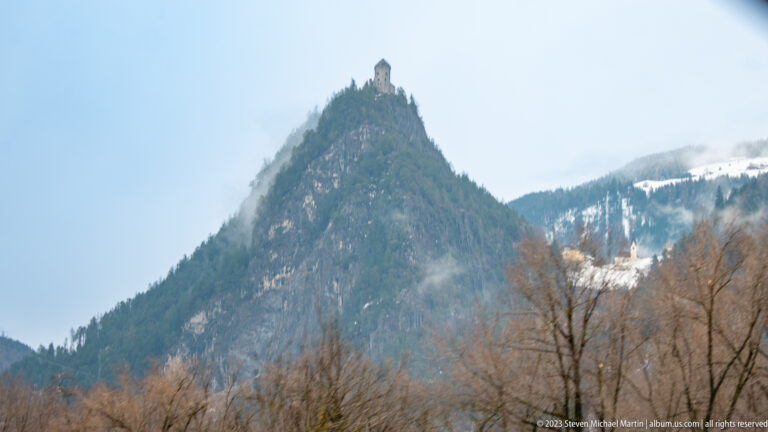 Small tower-like historic structure atop a steep conical wooded hill, with bare trees and snow in the valley below.