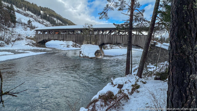 Long wooden covered bridge with open side trusses spanning a river between snowy banks and pine trees.