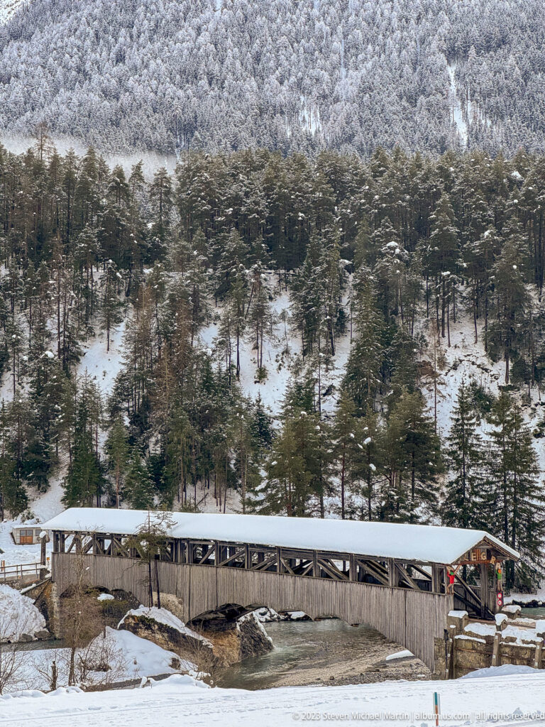 Broadside view of a long timber covered bridge with a snow-covered roof below a forested winter mountainside.