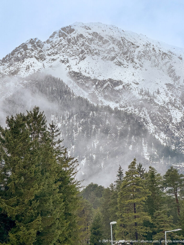 Snow-covered Alpine mountainside rising above dark evergreen forest, with mist or windblown snow drifting across the lower slopes.