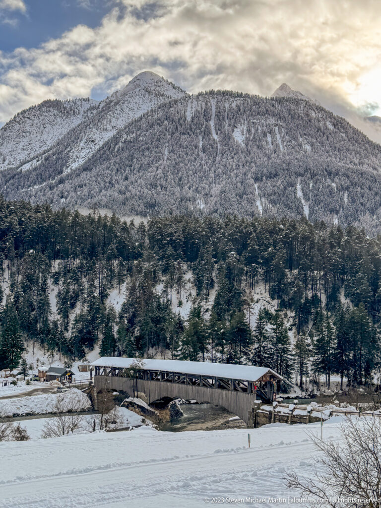 Long covered wooden bridge across a snowy valley with buildings and a broad forested mountain ridge behind.
