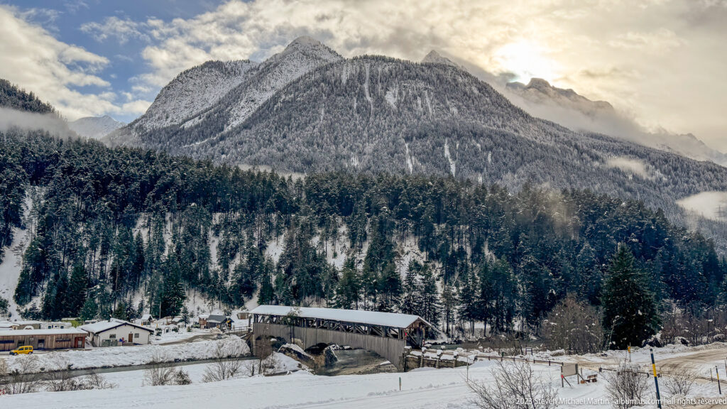 Snowy alpine valley with a timber-covered bridge, roadside buildings, dense conifer forest, and a steep mountain under broken clouds.