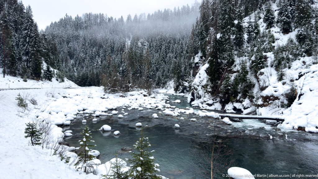 Dark mountain river winding between snowy banks, boulders, conifers, and misty forested slopes.