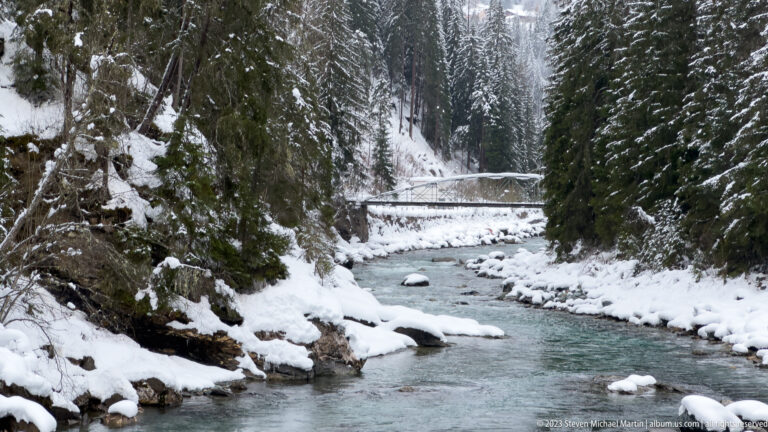 Blue-green river flowing between snowy conifer banks toward a narrow bridge in a steep valley.
