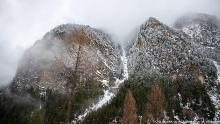 Jagged snow-dusted rock peaks above larch and evergreen forest, partly obscured by low cloud.