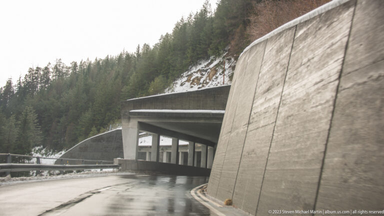 Wet mountain road curving beneath a large concrete protective gallery beside a snowy conifer slope.