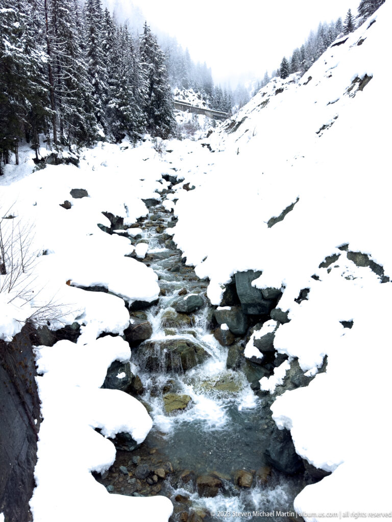 Shallow stream cascading over dark rocks through deep snow on an open alpine slope.