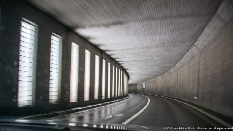 Curving wet roadway inside a concrete mountain gallery with tall vertical openings admitting daylight.
