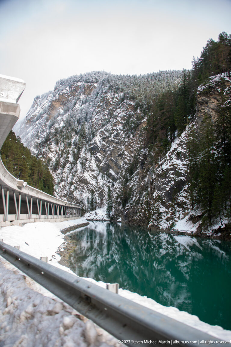 Turquoise mountain water beside a snowy road and steep conifer-covered slope.