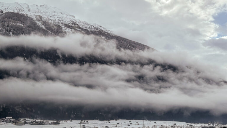 Low clouds band a dark forested mountain above a snowy valley floor with scattered buildings.