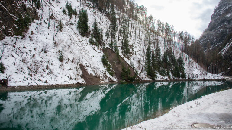 Still turquoise water reflecting a steep snowy hillside with conifers and bare larches.