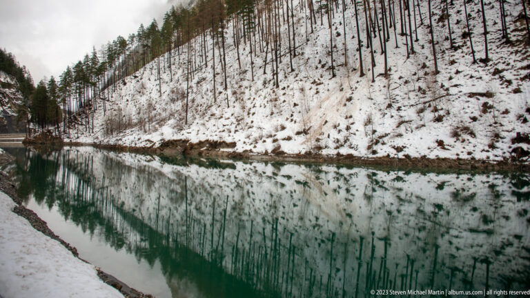 Calm green-blue water reflecting rows of trees on a steep snow-covered hillside.