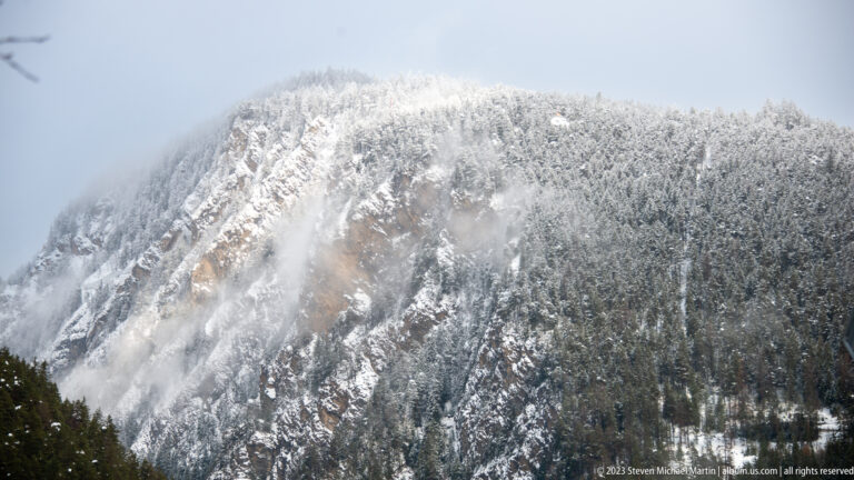 Snow-covered forested mountain wall with exposed rock and drifting mist obscuring the upper ridge.