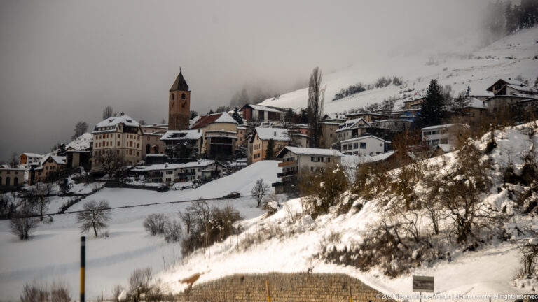 Snow-covered alpine village on a hillside with clustered houses and a prominent square church tower.