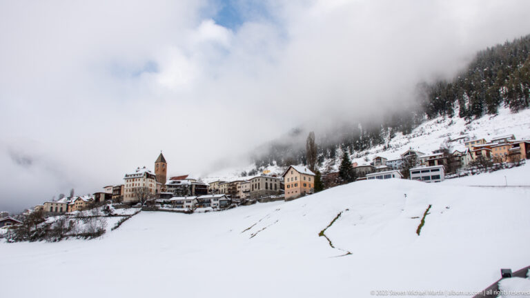 Wide snowy hillside with an alpine village, church tower, and forest disappearing into low cloud.