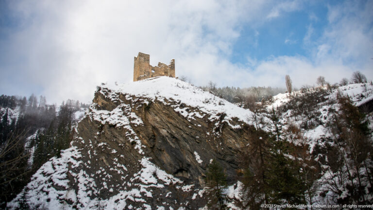 Broken stone tower ruin on a steep snow-covered rocky knoll above sparse winter vegetation.