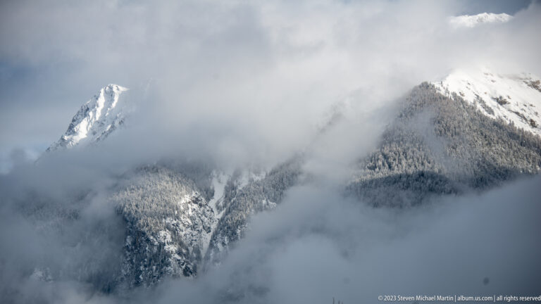 Sharp snowy mountain summit emerging from dense cloud above a darker forested ridge.