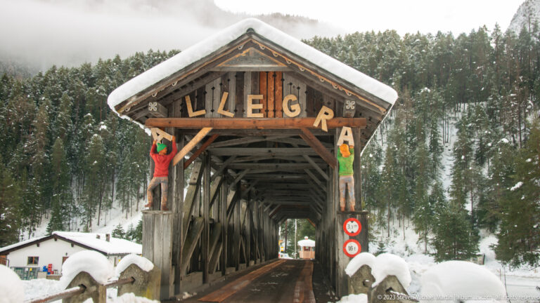 Front view of a timber covered bridge marked 'VAL S-CHARL,' with snowbanks, trusses, and road restriction signs.