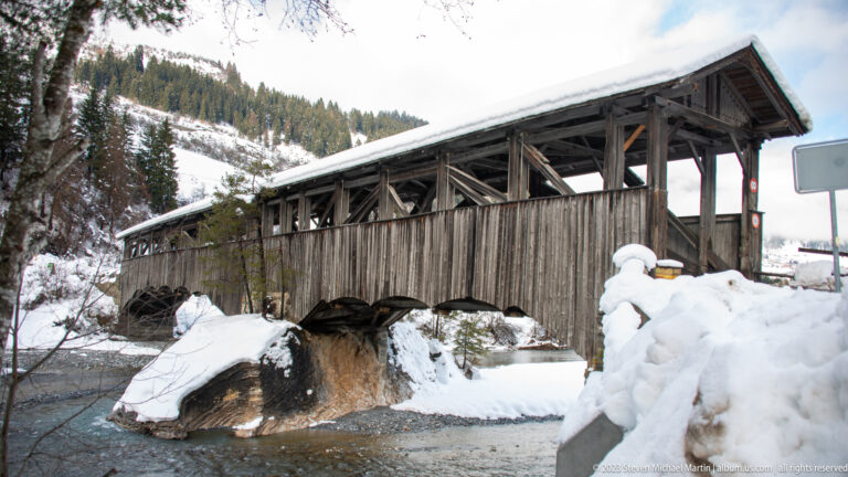 Weathered timber covered bridge spanning a rocky stream amid deep snow and forested mountain slopes.