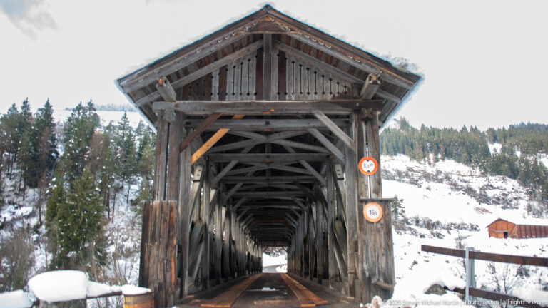 Centered view into a timber covered bridge with repeating trusses, snowbanks, forest, and restriction signs.