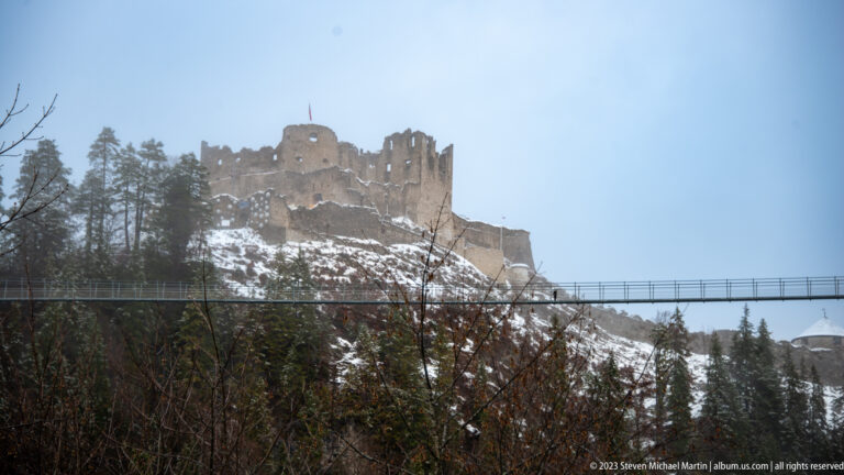 Roofless stone castle ruins on a snowy wooded hill above a long pedestrian bridge.