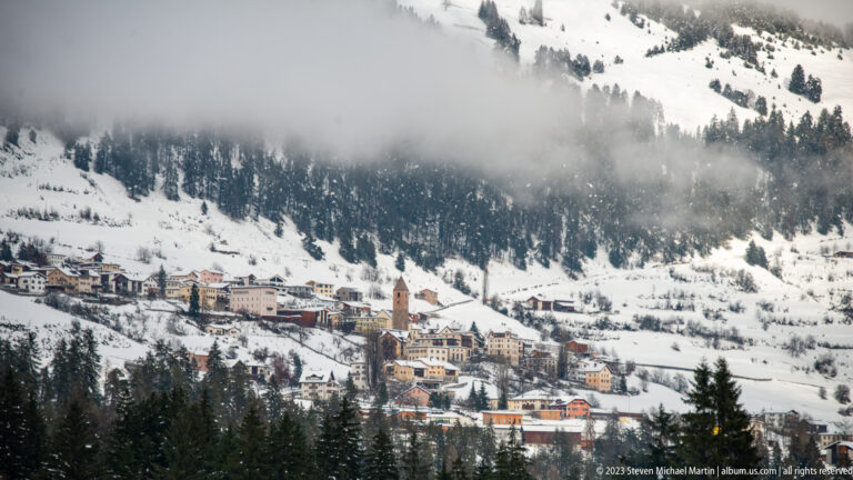 Elevated view across a snowy valley to an alpine village with a prominent church tower and forested slopes.