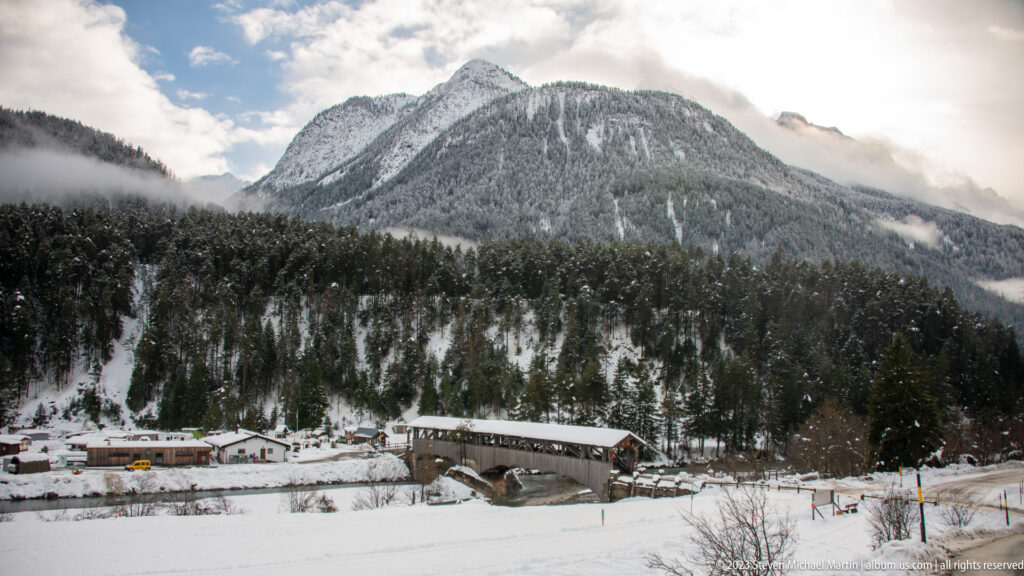 Snowy alpine valley with a timber-covered bridge, roadside buildings, forest, and a broad mountain under low cloud.