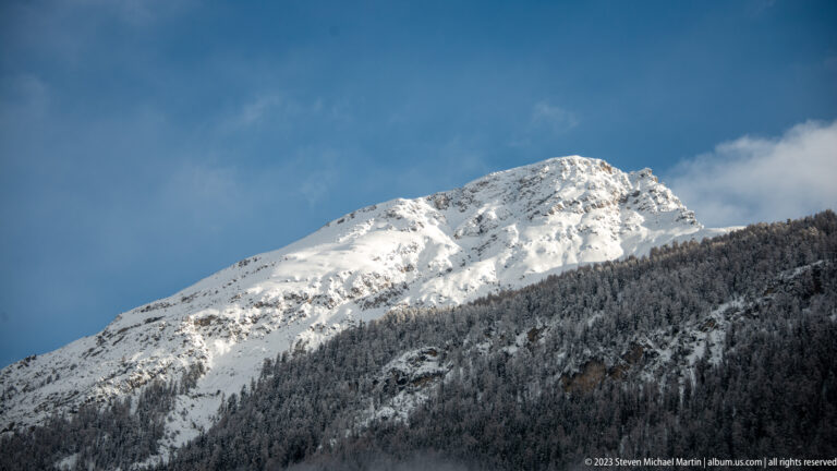 Sunlit snow-covered alpine ridge rising above dark forest beneath a deep blue patch of sky.