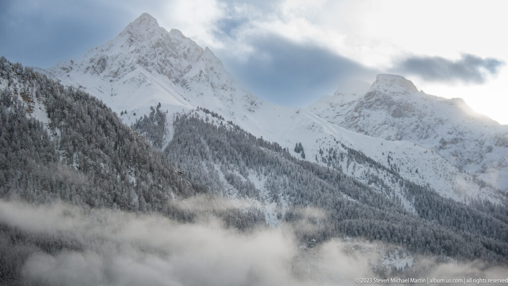 Snowy alpine ridges and a blocky rock peak above forested slopes and a valley filled with low cloud.