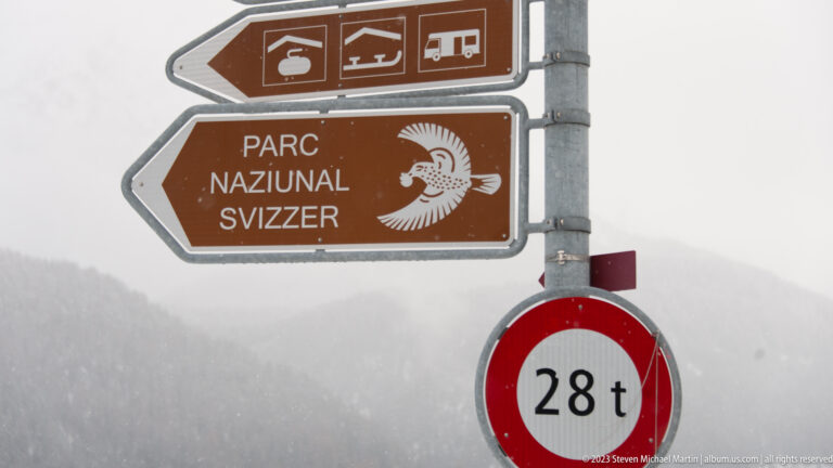 Brown 'Parc Naziunal Svizzer' directional sign with white bird emblem, tourist pictograms, and a 28-ton restriction sign.