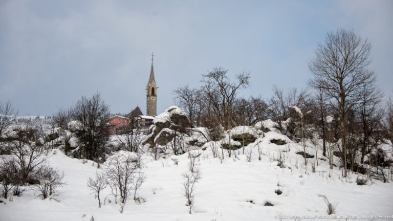 Slender church spire and red roof rising behind a snowy rocky hillside with bare trees.