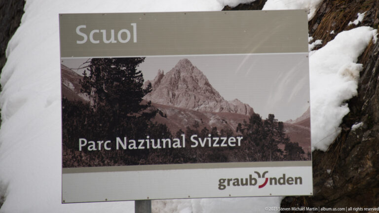 Roadside sign reading 'Parc Naziunal Svizzer' with a mountain photograph and graubünden logo beside snow-covered rocks.