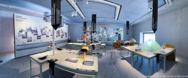 Modern visitor-center gallery with interactive tables, hanging labels, wall panels, models, and an illuminated topographic display.