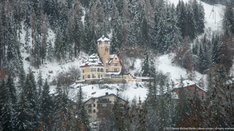 Ornate multi-story historic building with a tall square tower on a snowy conifer-covered hillside.