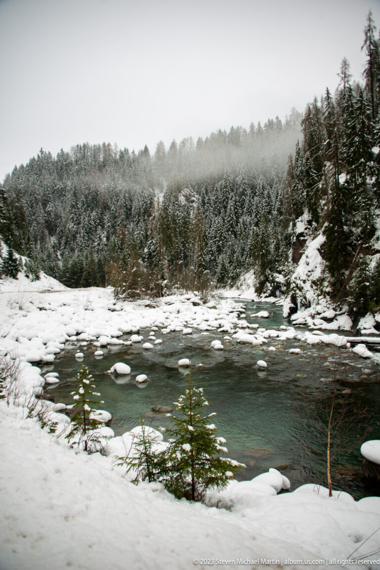 Broad dark-green river bend with snow-covered stones and banks beneath dense conifer forest.