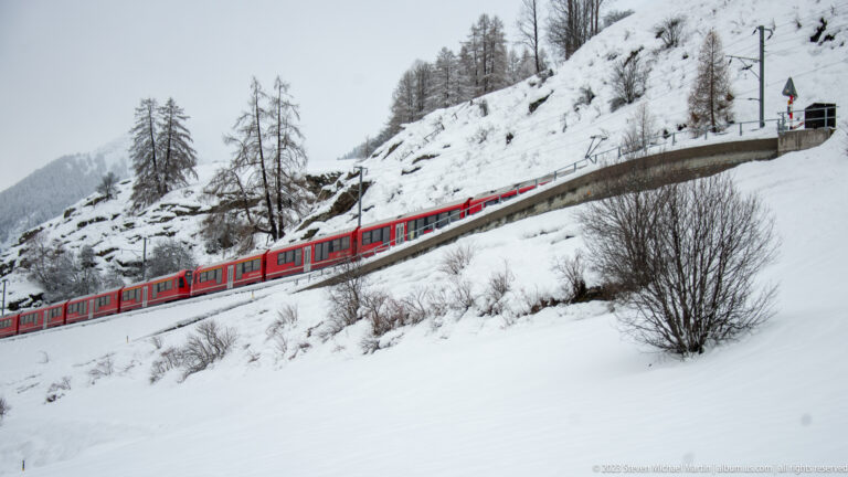 Long red passenger train traveling uphill on an electrified railway across a snowy alpine slope.