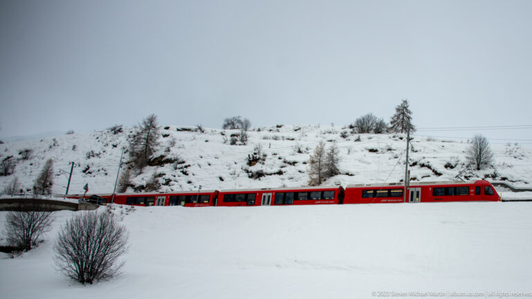Red electric passenger train in full side profile crossing a snow-covered hillside beneath catenary wires.