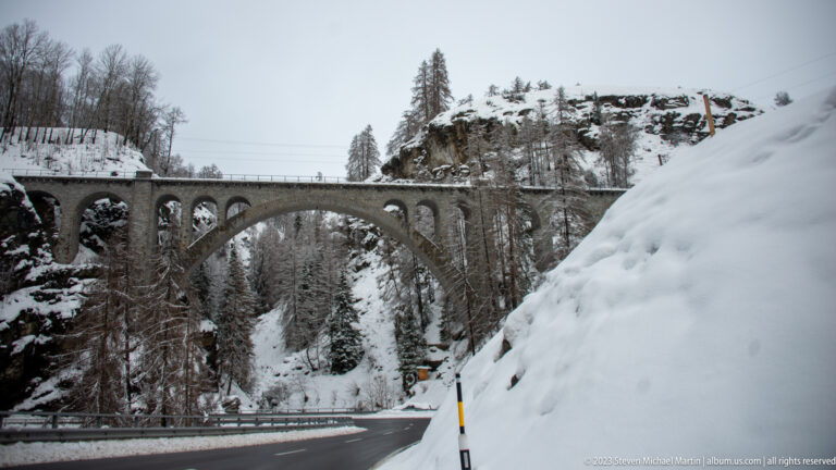 Large multi-arched stone railway viaduct spanning a snowy ravine above a curving road.