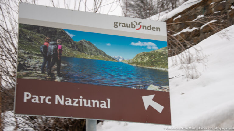 Brown graubünden tourism sign reading 'Parc Naziunal' with a left arrow and mountain-lake photograph.