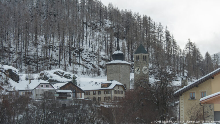Snowy alpine village with a stone clock tower, second square tower, houses, and larch-covered slope.