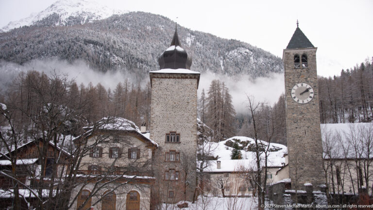 Paired stone towers, one with a clock and one with an onion-shaped roof, in a snowy alpine village.