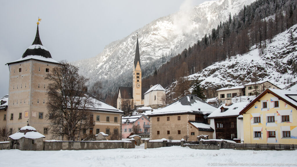 Snowy alpine village with a broad square tower, slender church spire, clustered houses, and steep mountains.