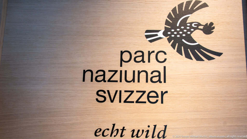Wood-toned panel with the Parc Naziunal Svizzer bird logo, lowercase name, and 'echt wild' slogan.