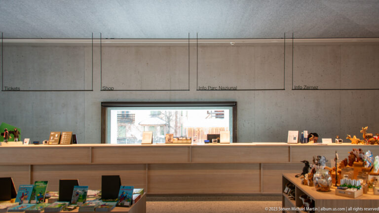 Wooden visitor-center counter with books, souvenirs, display items, and wall labels for tickets, shop, and information.