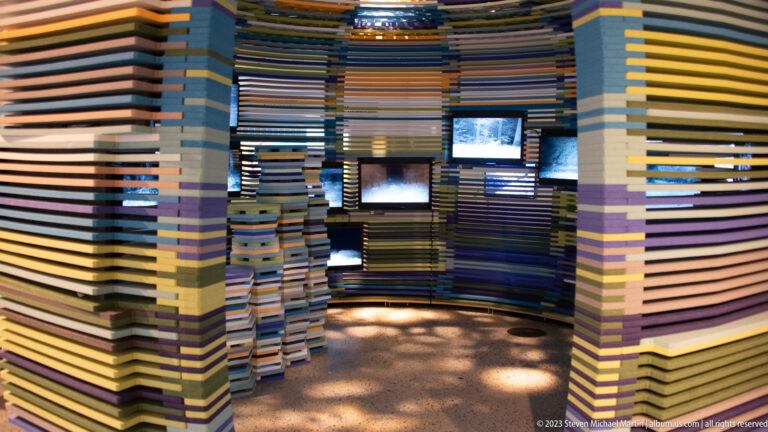Immersive gallery with stacked multicolored slat walls, small screens, niches, and pools of floor lighting.