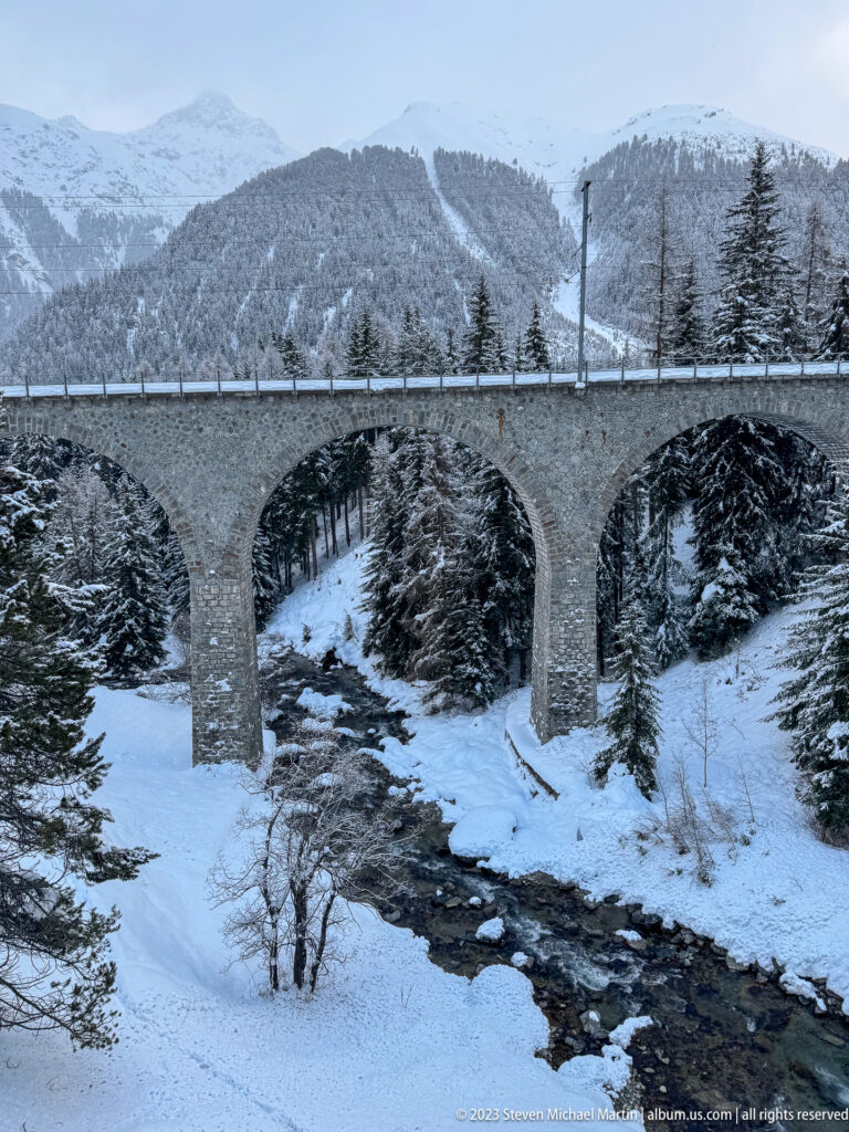 Multi-arched stone viaduct crossing a dark mountain stream through snow-covered conifer terrain.