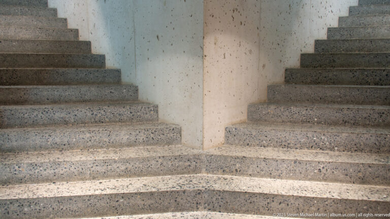 Symmetrical concrete stair flights rising around a central wall, with speckled treads and slim handrails.
