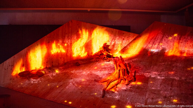 Dark gallery with projected flames across angled walls and a charred root or trunk installation on the floor.