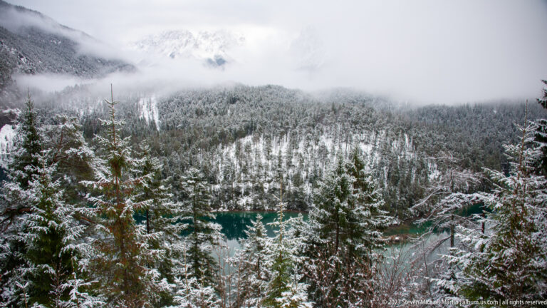 Turquoise Alpine lake partly visible through snow-covered evergreen trees beneath misty slopes.